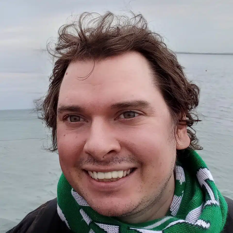 Homem sorridente com cabelo despenteado junto a um mar calmo